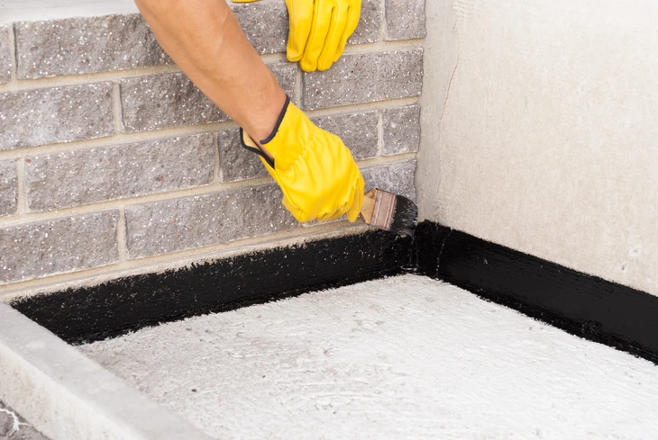 Personne appliquant un produit d'étanchéité noir au pinceau le long du joint entre mur en briques et surface blanche, portant des gants jaunes de protection