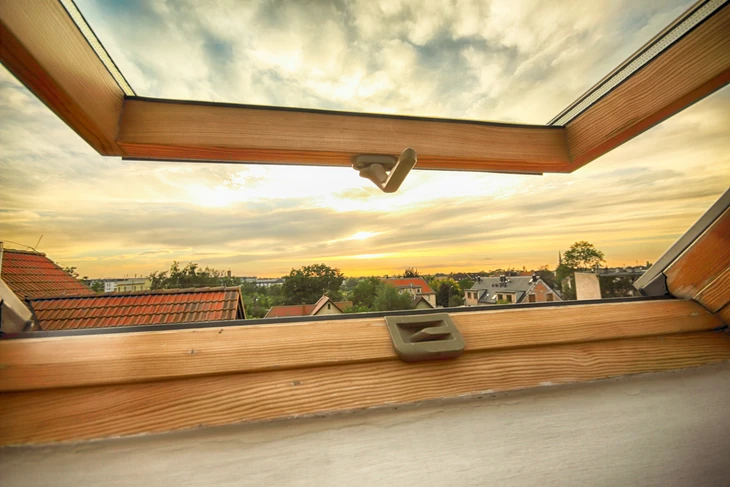 Fenêtre de toit ouverte dans combles aménagés révélant vue panoramique sur toitures résidentielles au coucher de soleil
