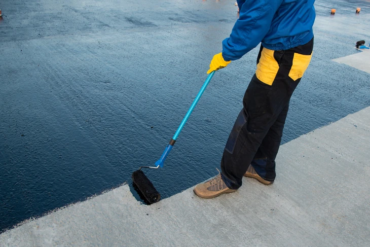 Ouvrier en combinaison bleue et jaune applique un revêtement d'étanchéité bleu sur surface béton extérieure avec rouleau à manche télescopique