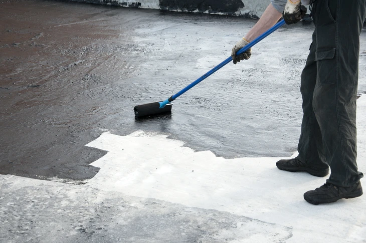 Ouvrier appliquant un revêtement d'étanchéité liquide sur une toiture-terrasse en béton à l'aide d'un rouleau à manche télescopique bleu