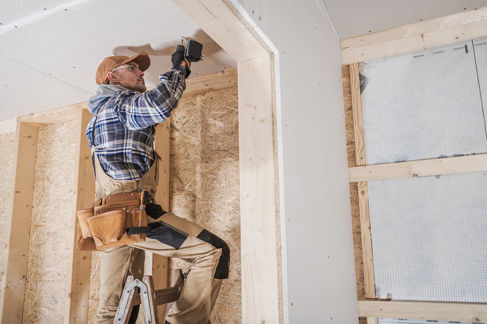 Ouvrier installant un système électrique dans une construction en bois, travaillant depuis un escabeau dans un espace intérieur avec ossature bois et isolation
