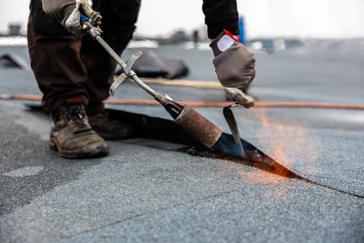 toit professionnel appliquant un rouleau de bitume sur un toit plat avec brûleur à gaz sur un chantier de construction moderne