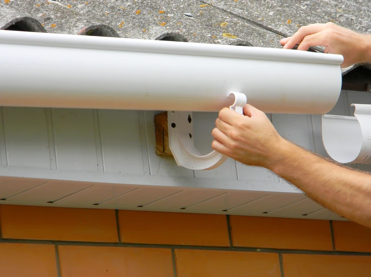 Installateur posant une gouttière blanche en PVC sur bordure de toit avec mur en briques oranges