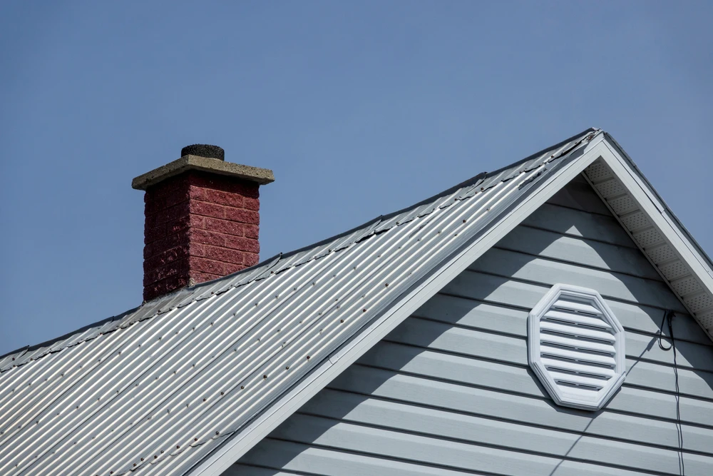 Extérieur résidentiel avec toiture en tôle ondulée métallique grise, cheminée en brique rouge, pignon à bardage vinyle horizontal bleu-gris avec ventilation circulaire blanche