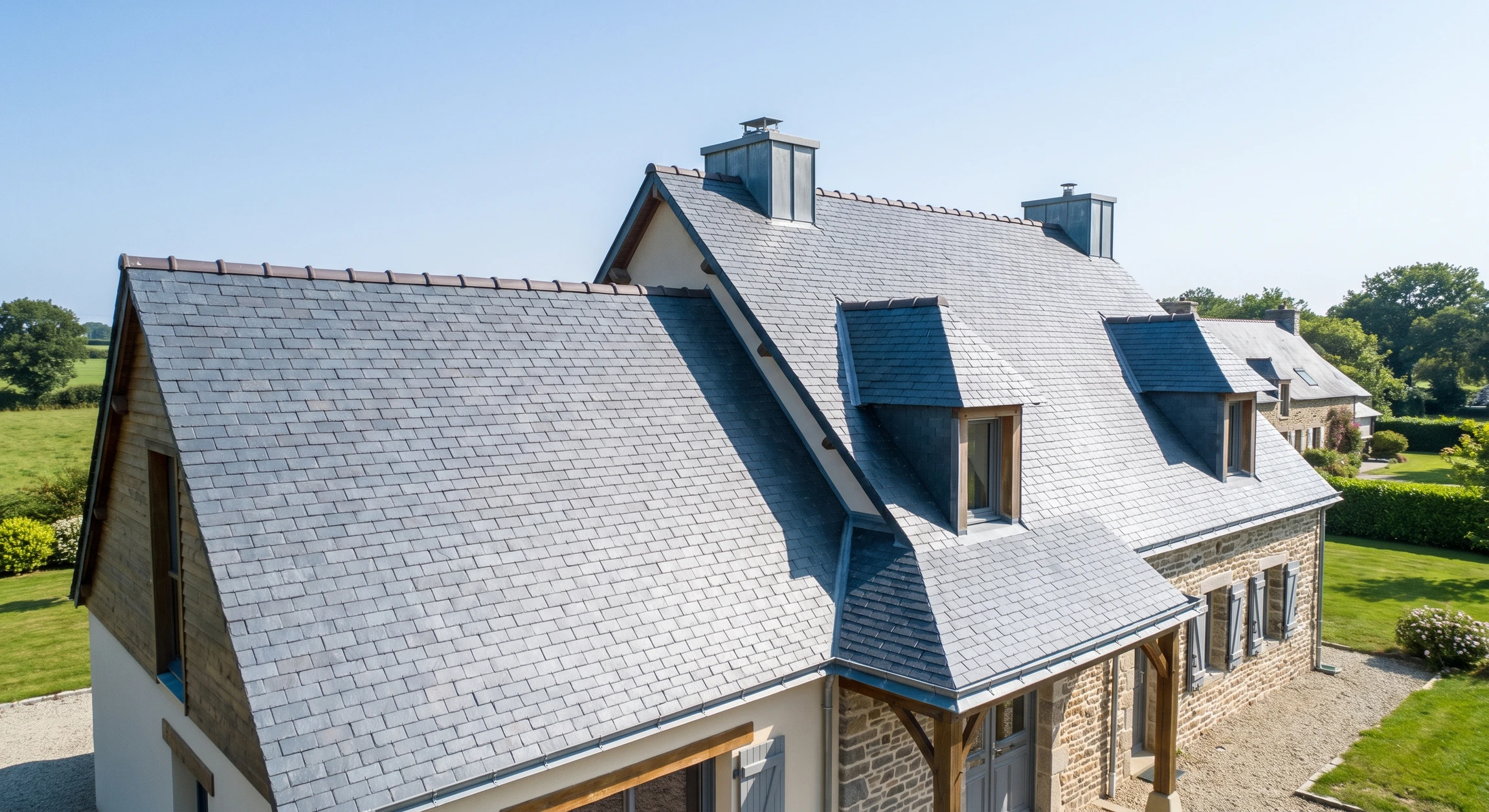 Photographie d'une maison résidentielle de caractère avec toit entièrement couvert d'ardoise naturelle, montrant clairement les tuiles d'ardoise gris-bleuté dans toute leur texture et leur brillance, avec pentes et détails de zinguerie visible. L'angle de prise de vue permet d'apprécier la qualité et l'élégance de la pose, le contexte résidentiel renforce l'message "habitat de caractère".