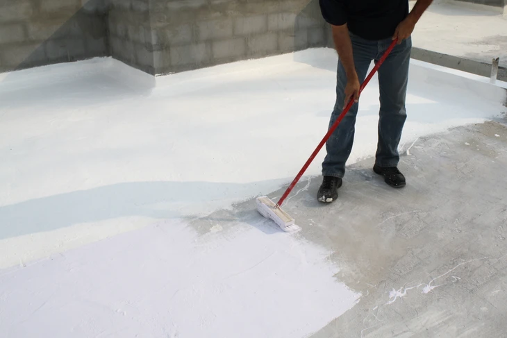 Homme appliquant un revêtement d'étanchéité blanc sur une toiture-terrasse en béton avec un rouleau à manche rouge