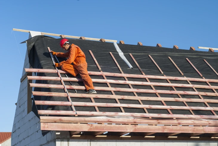 Ouvrier en combinaison orange et casque rouge installe une membrane d'étanchéité noire sur charpente en construction