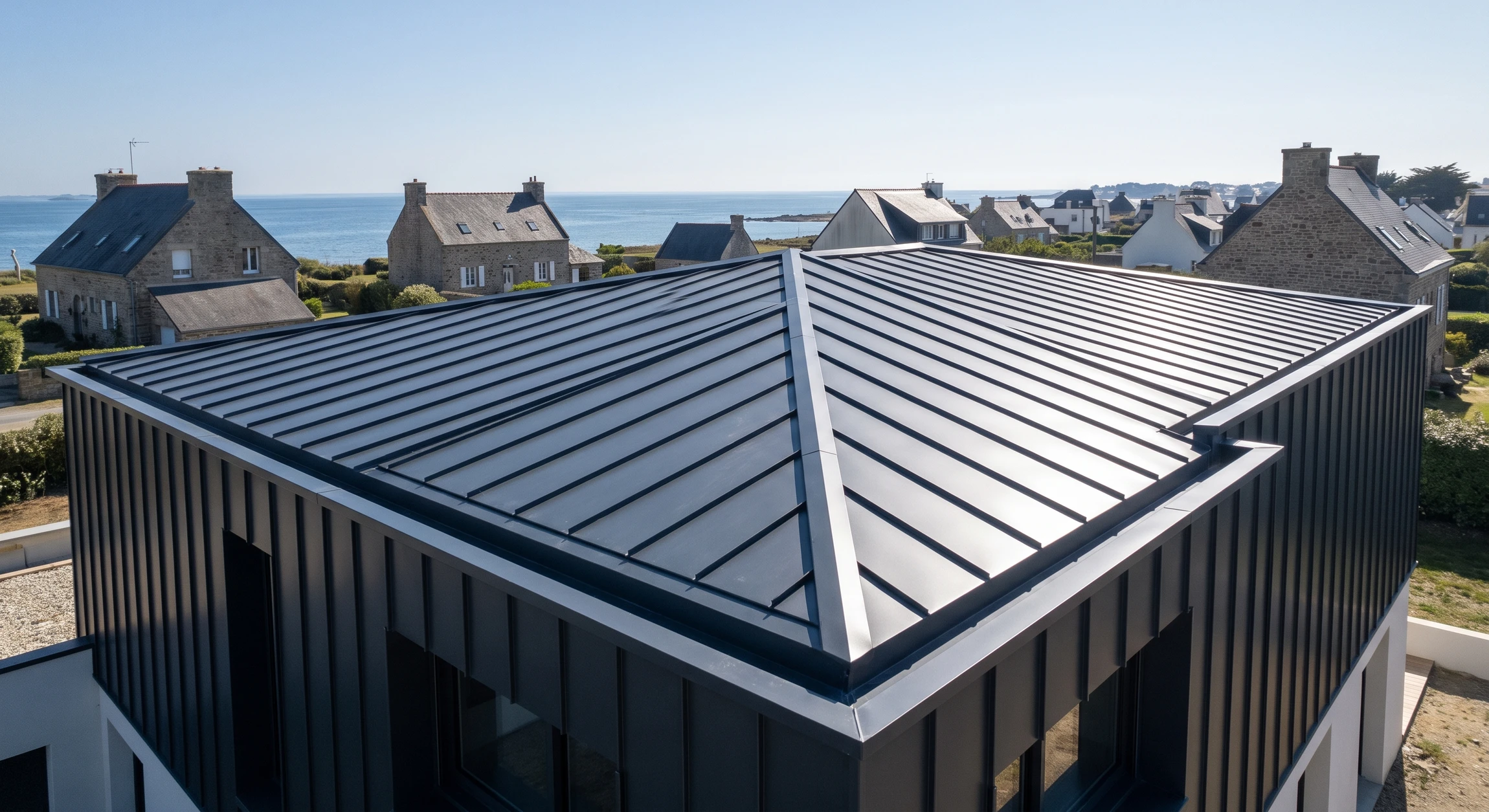Photographie en plongée ou trois-quarts d'une toiture résidentielle ou commerciale couverte de panneaux ou tôles d'aluminium brillant ou gris anthracite, avec des détails de zinguerie d'aluminium visibles (noues, arêtes, gouttières). La toiture doit être bien exposée sous un éclairage naturel mettant en valeur la finition métallique et la qualité de la pose. Aucun texte, logo ou filigrane visible.