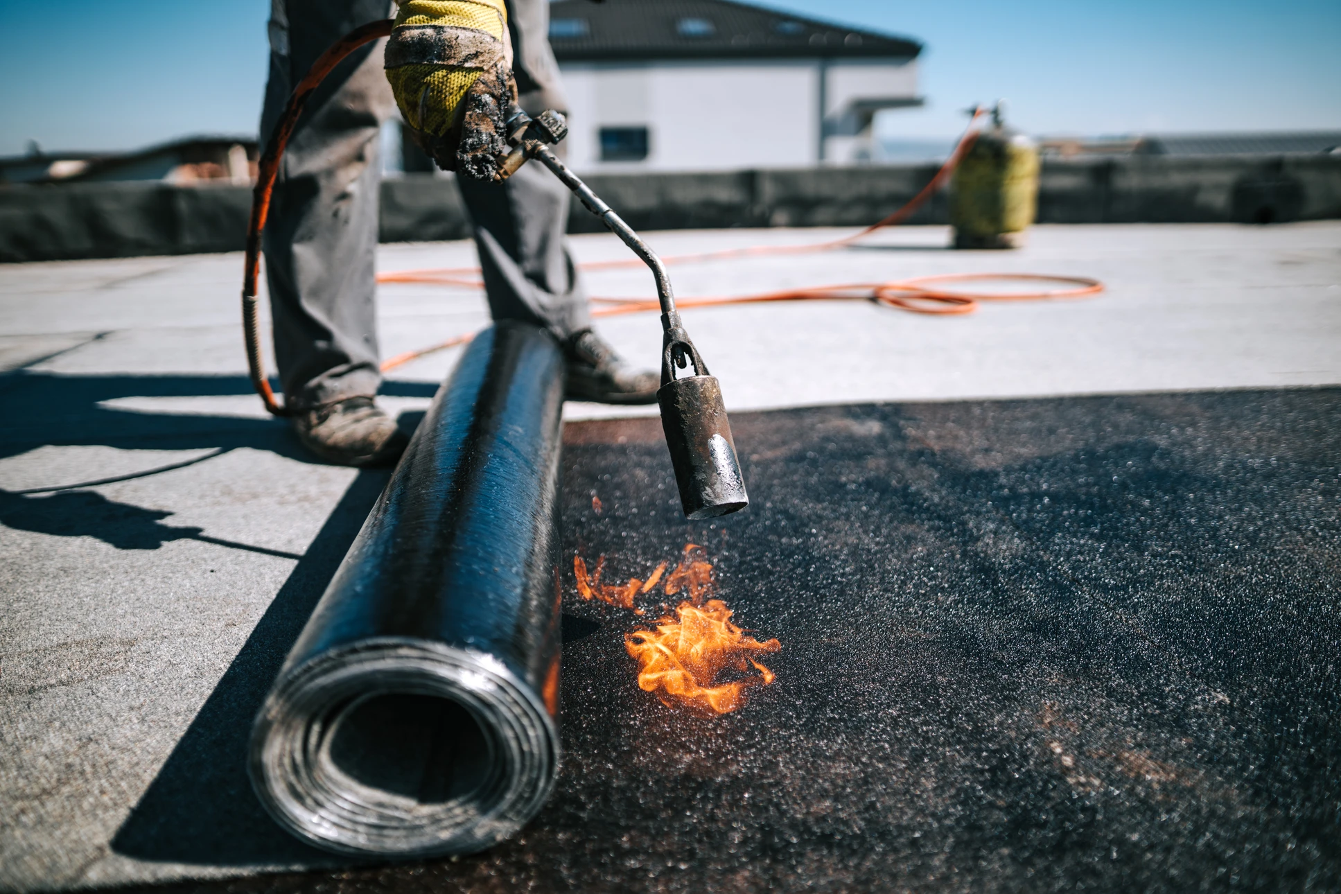 Photographie d'un professionnel en tenue de chantier effectuant des travaux d'étanchéité sur un toit terrasse ou une toiture plate, montrant la mise en place de membrane d'étanchéité ou l'application de résine protectrice. L'image doit illustrer le savoir-faire technique face à des conditions climatiques côtières (ciel gris, exposition maritime). Vue en plongée ou plan large montrant l'ampleur des travaux et la qualité professionnelle de l'intervention.